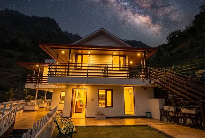 Madhuram Stays Resort exterior at dusk — warm lights, mountain backdrop